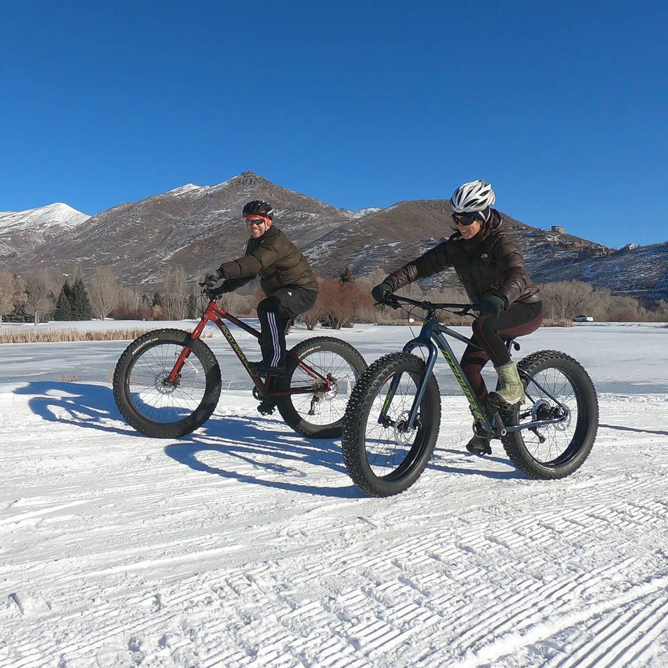 Featured Contributor, Jessica Averett (right), wears a winter jacket, sunglasses, and white bike helmet while riding a Fat Tire bike in the snow next to a man in front of the mountains.