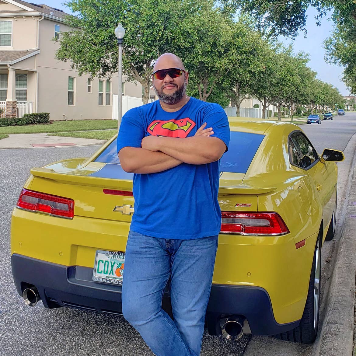 A man in a blue, t-shirt with the Superman emblem wears sunglasses and jeans while leaning against a yellow Camaro on a street.