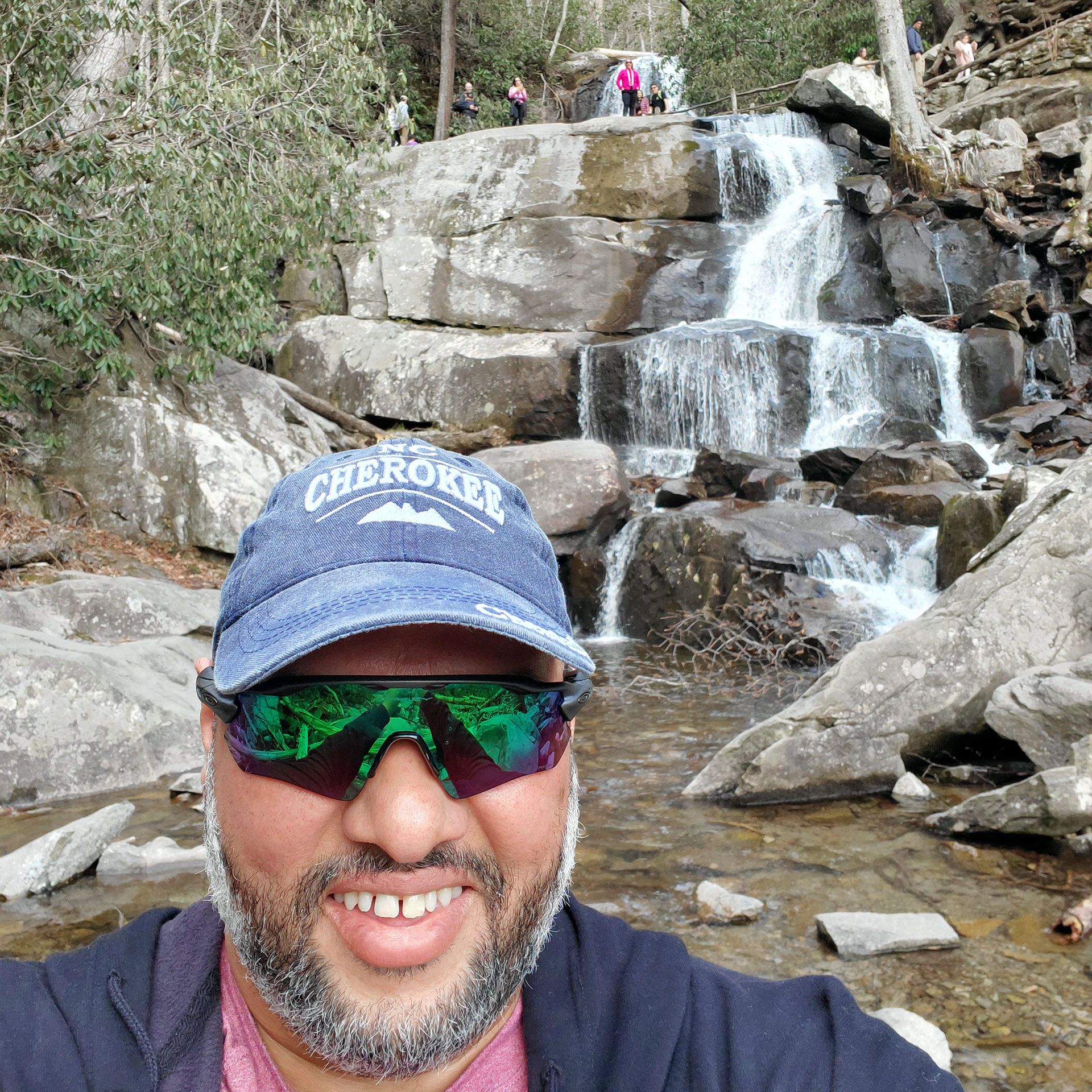 A man wearing a denim baseball cap that reads, 'Cherokee', sunglasses and a navy zip-up hoodie stands in front of a waterfall feature in a forest.