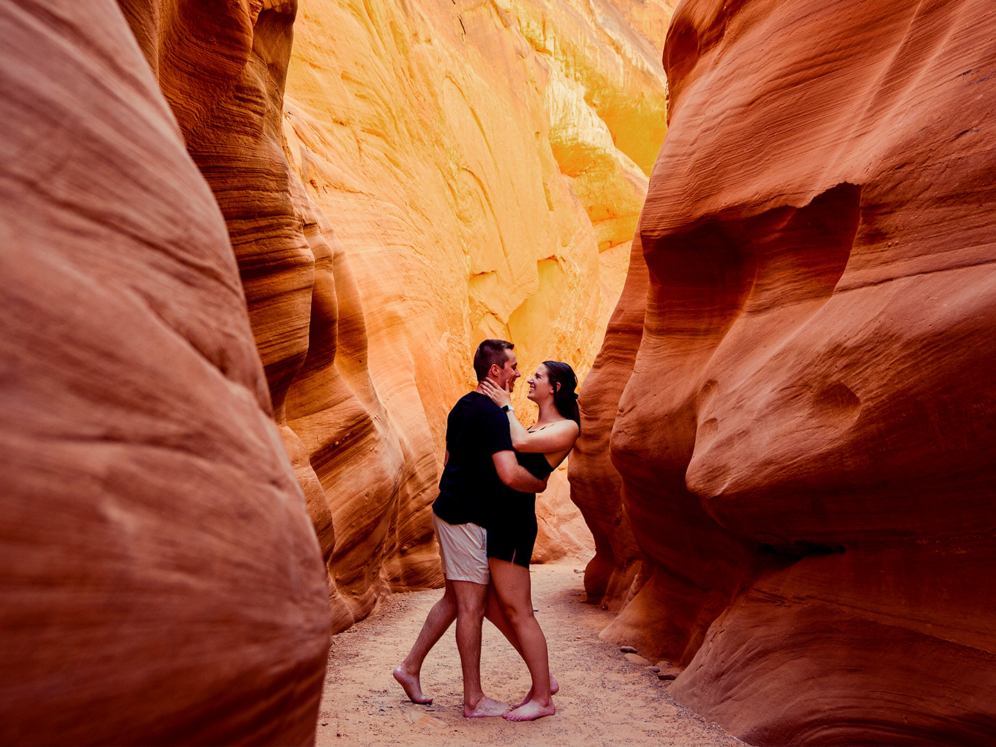 A man and woman wearing black outfits embrace in a red rock formation.