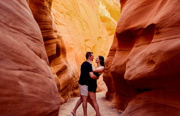 A man and woman wearing black outfits embrace in a red rock formation.