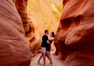 A man and woman wearing black outfits embrace in a red rock formation.
