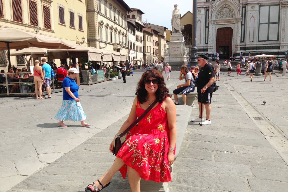Author, Jennifer Probst, sits on a bench in the middle of Venice wearing a red sundress and sunglasses with a black bag slung across her shoulder.