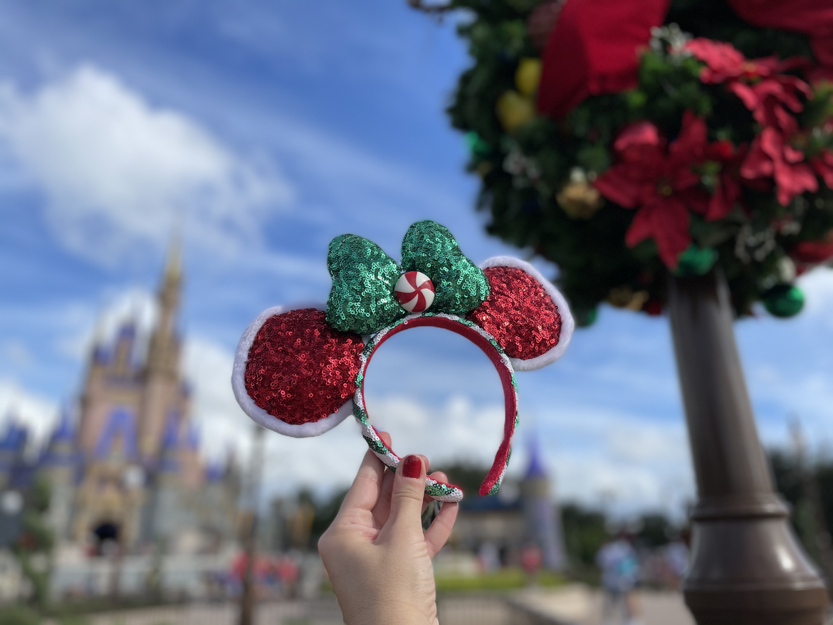 Author, Kelly Nelson, holds a pair of red sequin, white-fuzz-lined Minnie Mouse Ears with a large green bow centered with a peppermint candy in front of Cinderella's Castle located at the Magic Kingdom Park at Walt Disney World® Resort.