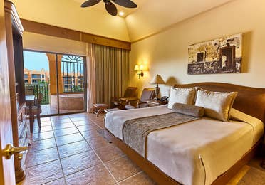 King size bed, covered in a brown and golden spread with 3 golden decorative pillows, a seating area with 2 chairs and an angled view of the tiled jacuzzi on the balcony at The Royal Haciendas in Playa del Carmen, Mexico