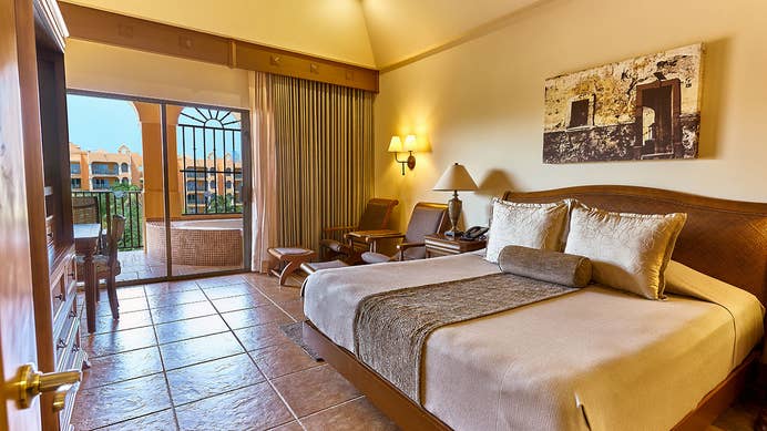 King size bed, covered in a brown and golden spread with 3 golden decorative pillows, a seating area with 2 chairs and an angled view of the tiled jacuzzi on the balcony at The Royal Haciendas in Playa del Carmen, Mexico