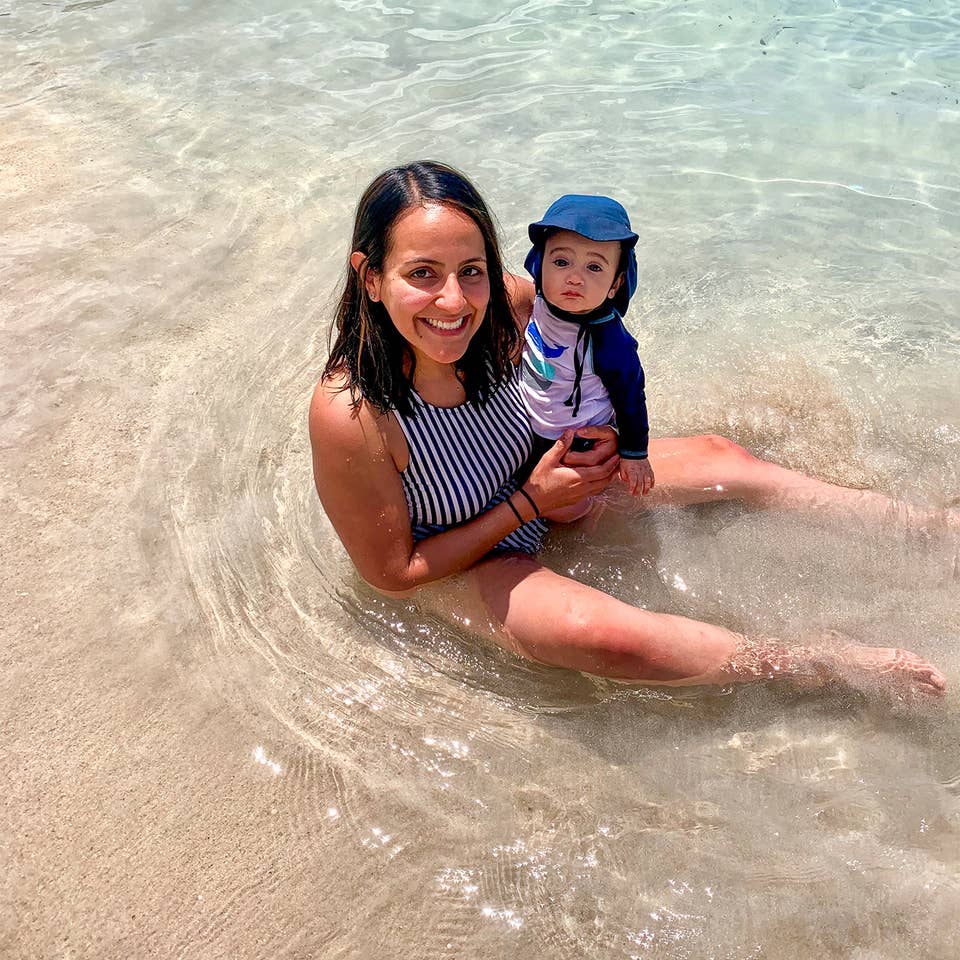 A woman in a striped swimsuit holds a baby wearing swimsuit and a sunhat in shallow water.