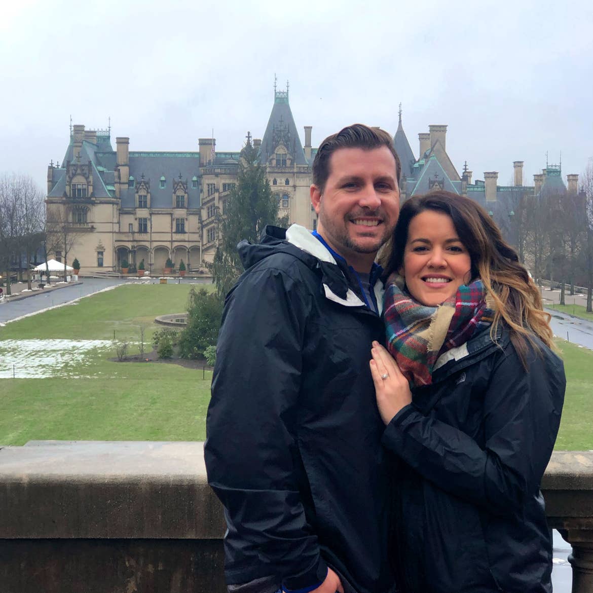 Author, Jenn C. Harmon (right), and husband (left) stand in front of the Biltmore Estate wearing black jackets and a scarf on a grey cloudy day.