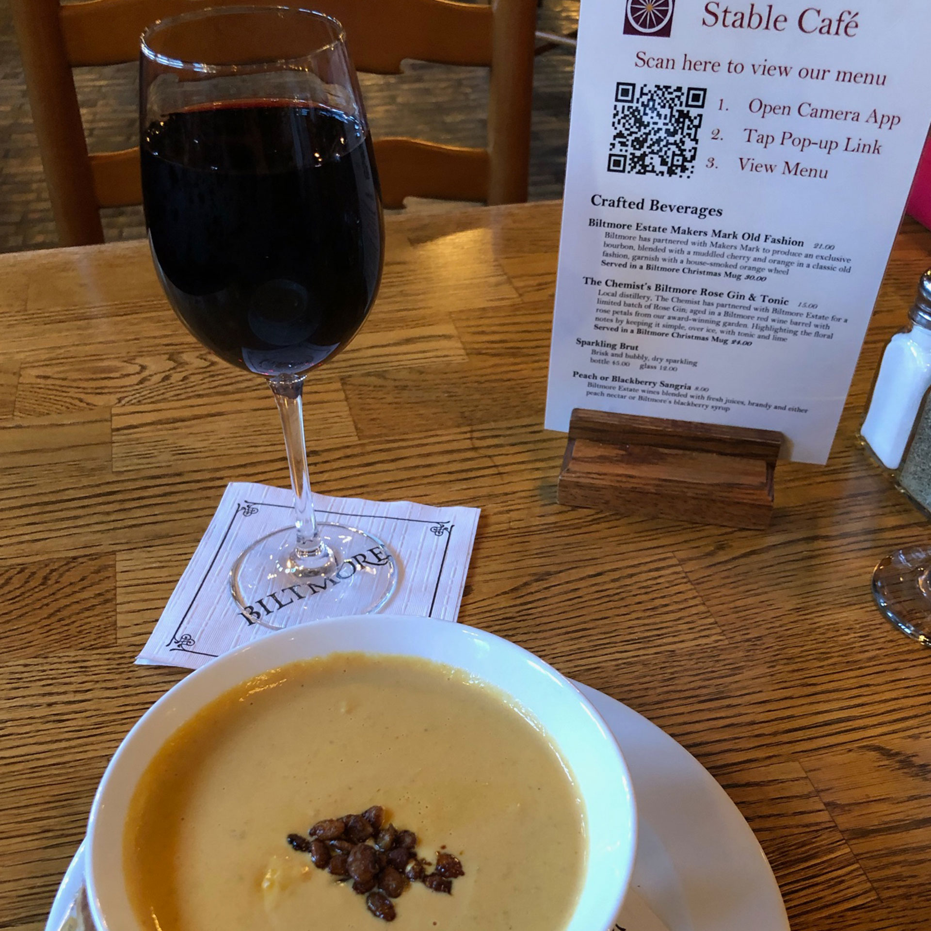 A glass of red wine and the pumpkin bisque are placed on a table near the QR menu at the Stable Cafe.