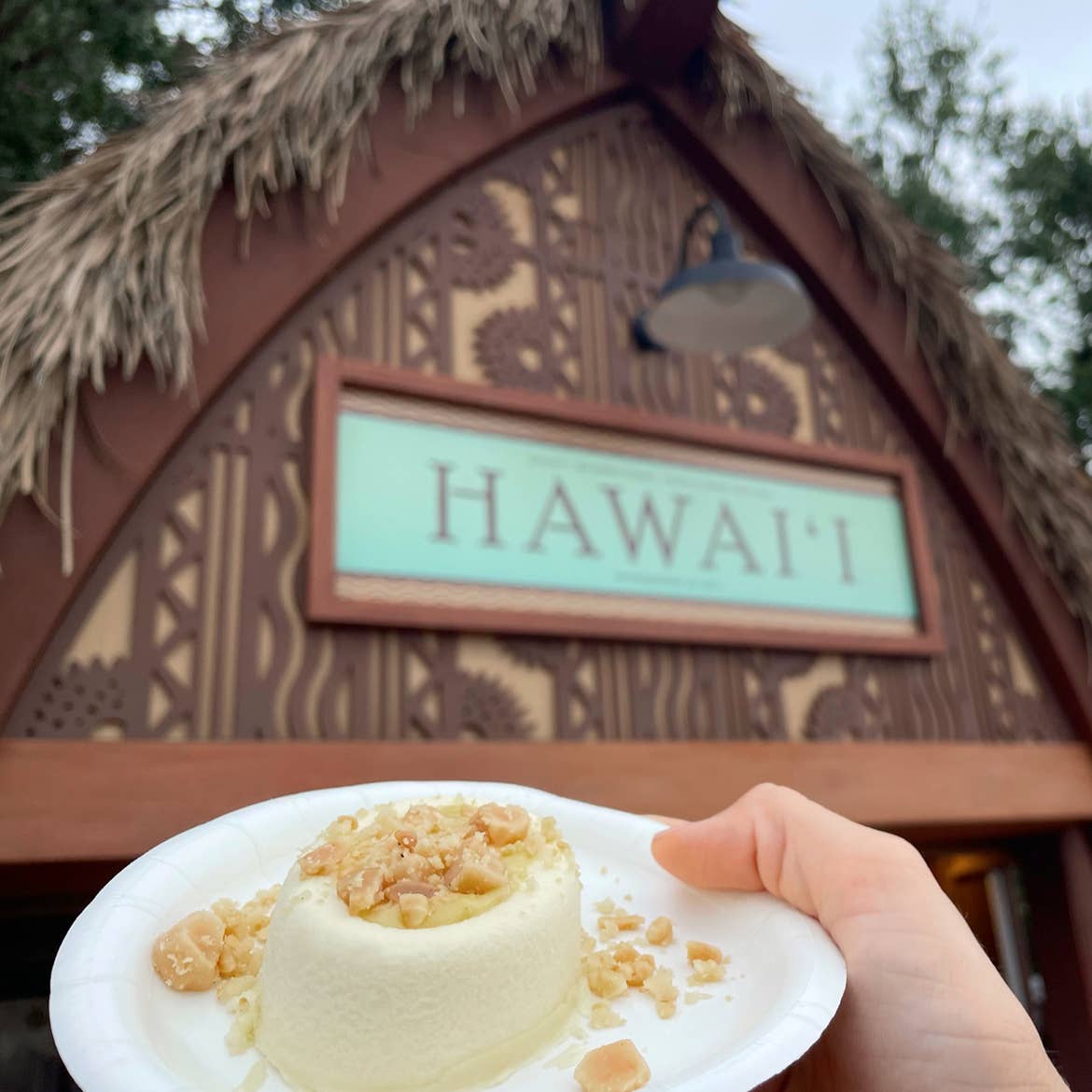 A hand holds a plate containing a Passionfruit Cheesecake with Toasted Macadamia Nuts near the Hawaii kiosk at Epcot.