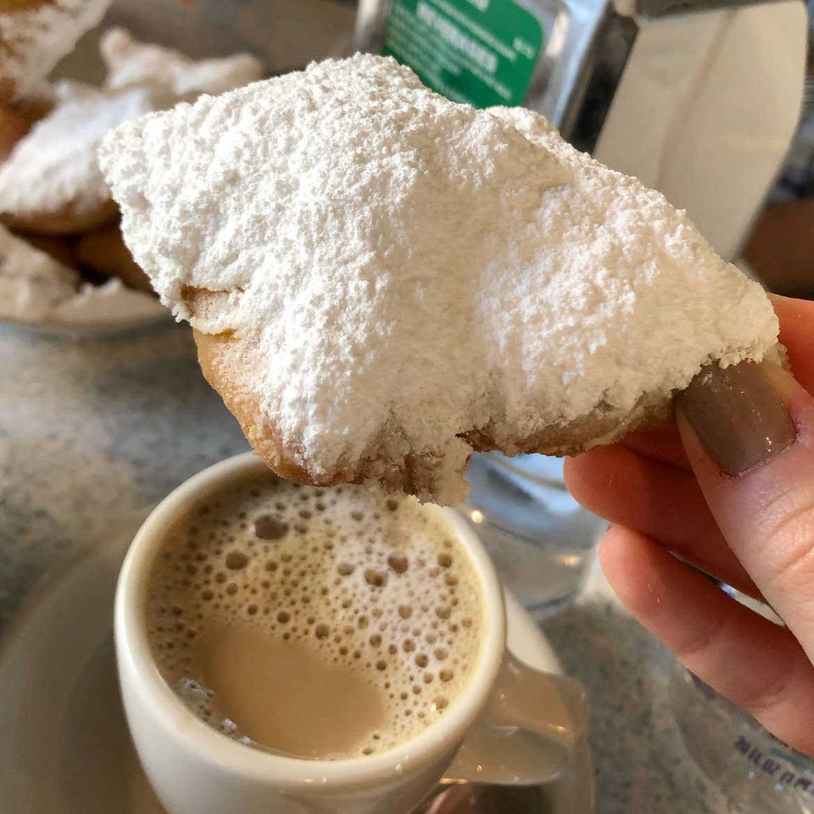 A Cafe Du Monde napkin box and a white coffee mug placed on a table while a beignet is held.