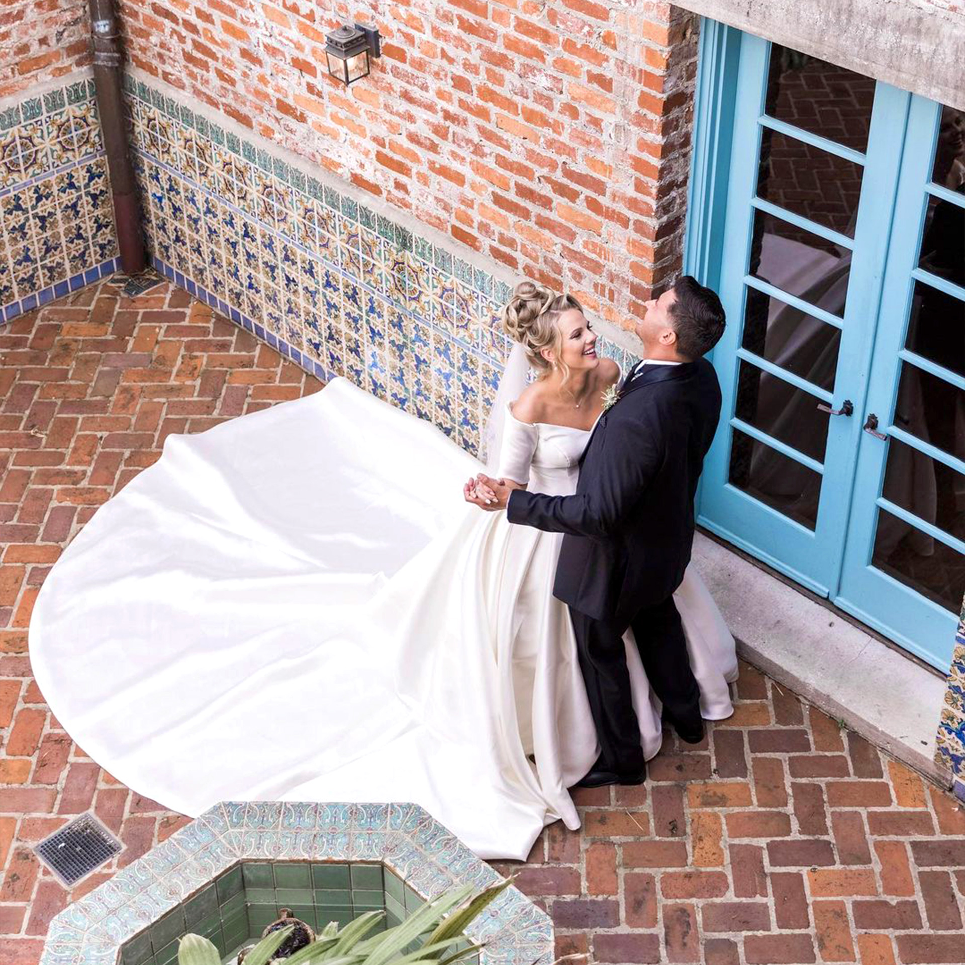 A Latinx groom wearing a black suit and (right) and a caucasian bride with a long train (left) dance with each other in front of a brick facade.
