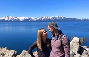 A woman (left) black pullover while hugging a man (right) wearing a red pullover and grey hat in front of a mountain range at Lake Tahoe, Nevada.