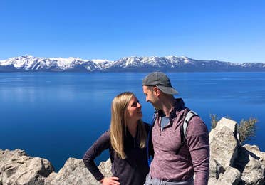A woman (left) black pullover while hugging a man (right) wearing a red pullover and grey hat in front of a mountain range at Lake Tahoe, Nevada.