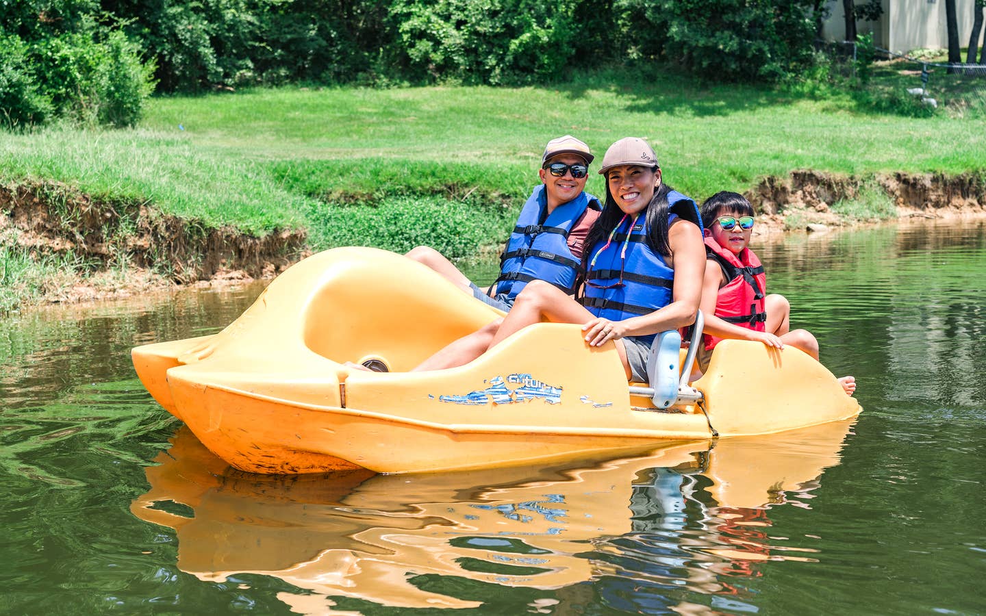 An Asian Male (left), female (middle) and young child (right) wear blue life vests, sunglasses and various hats while riding on a yellow paddleboat in a lake.