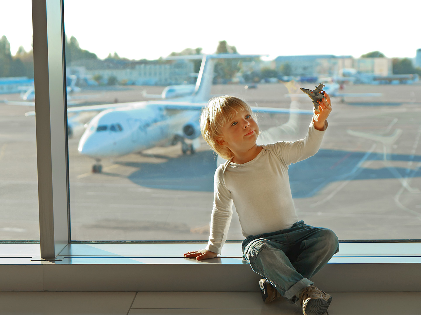 A young boy wearing a white long sleeve shirt plays with a toy airplane in front of an airport window near the runway with a white airplane.