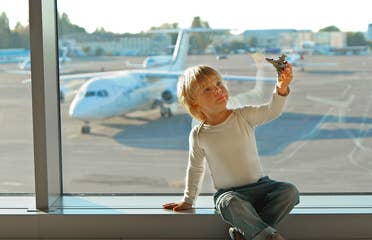A young boy wearing a white long sleeve shirt plays with a toy airplane in front of an airport window near the runway with a white airplane.