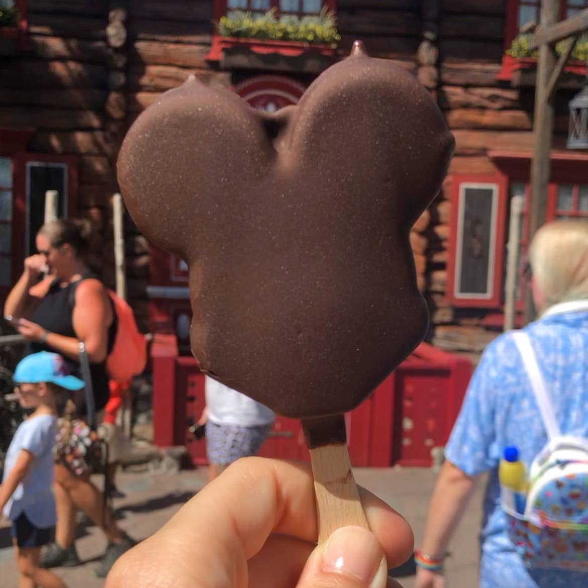 A hand holds a Mickey Ice Cream bar in front of the Royal Sommerhus at the Norway Pavilion in Epcot.