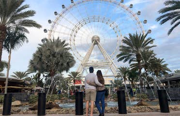 Featured contributor, Tori Ferrante (right) and her husband embrace in front of the Orlando Icon Ferris Wheel at ICON park under a blue sky.