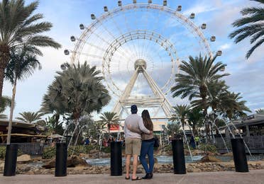 Featured contributor, Tori Ferrante (right) and her husband embrace in front of the Orlando Icon Ferris Wheel at ICON park under a blue sky.