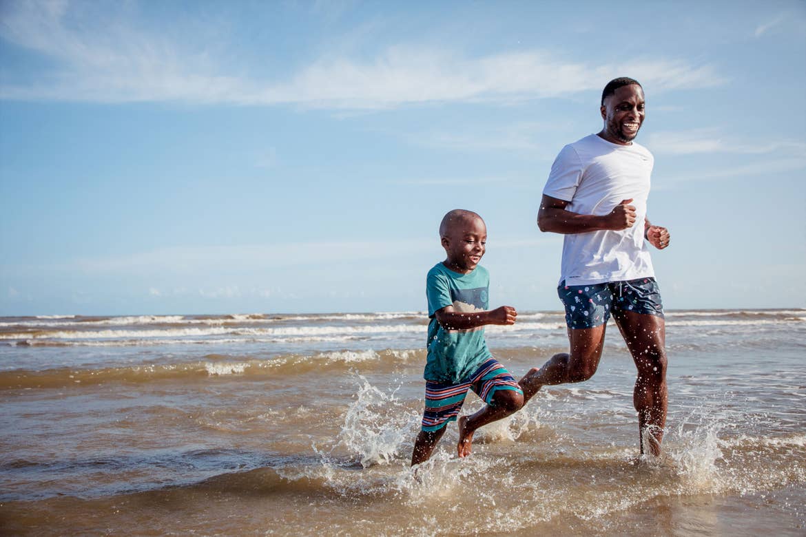 A man and young boy run through water on a beach.