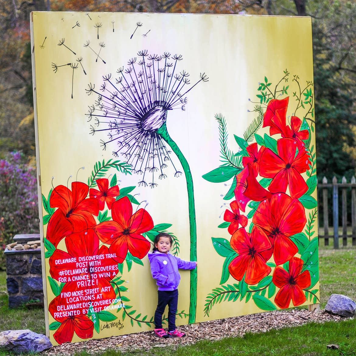 A young girl in a purple coat poses near a mural as if she were holding a purple dandelion surrounded by red poppy flowers outdoors.