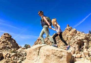 A woman and child hike across red rock formations under a blue sky.