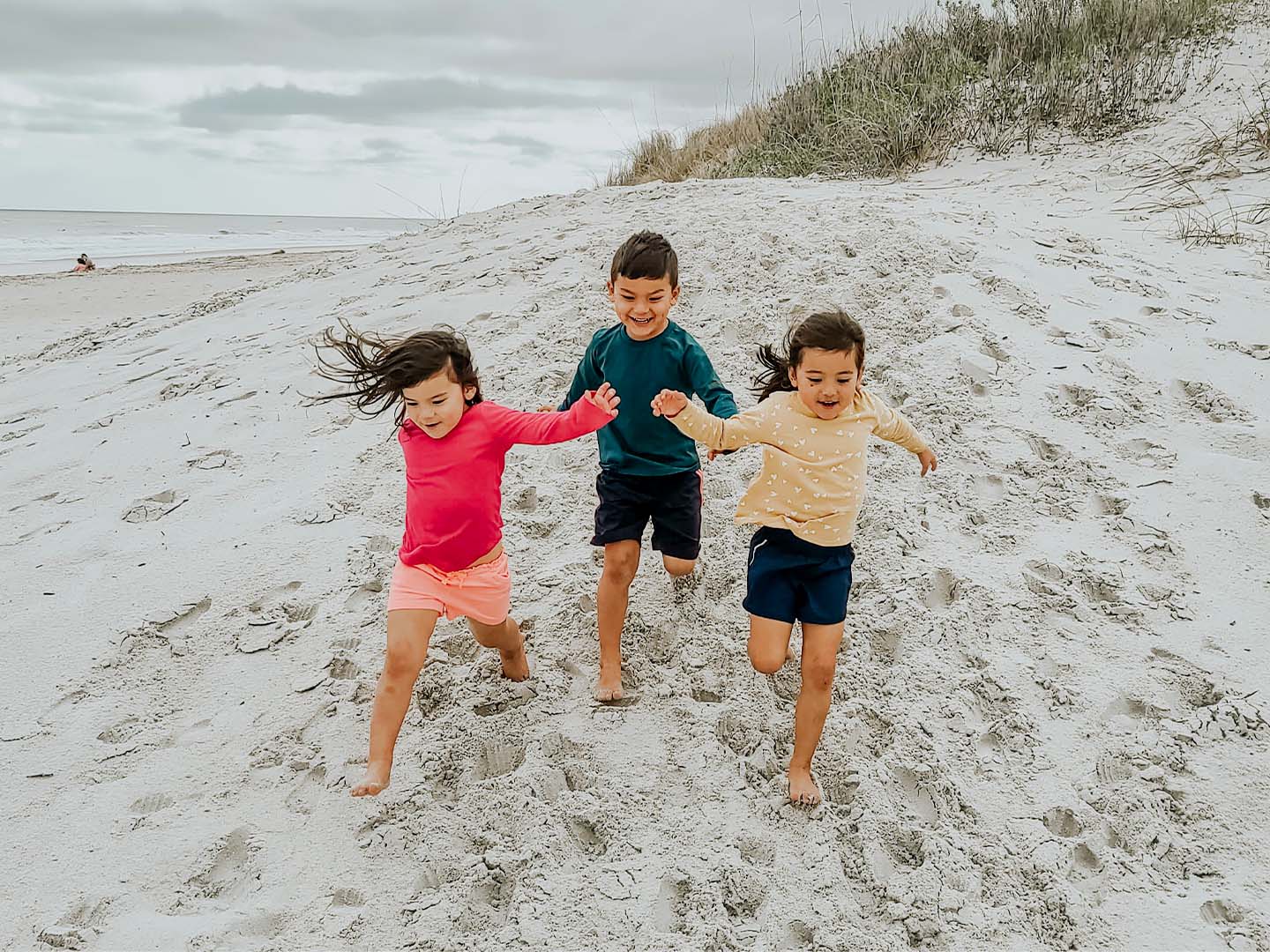 Three toddlers in multi-colored rash guards and board shorts run on a sandy beach.