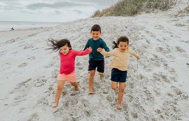 Three toddlers in multi-colored rash guards and board shorts run on a sandy beach.