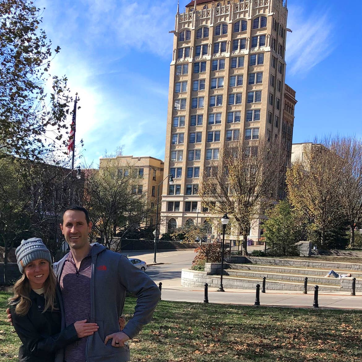 A woman (left) wearing a black jacket and grey beanie while hugging a man (right) wearing a red pullover and grey backpack in front of a building at downtown Asheville, North Carolina.
