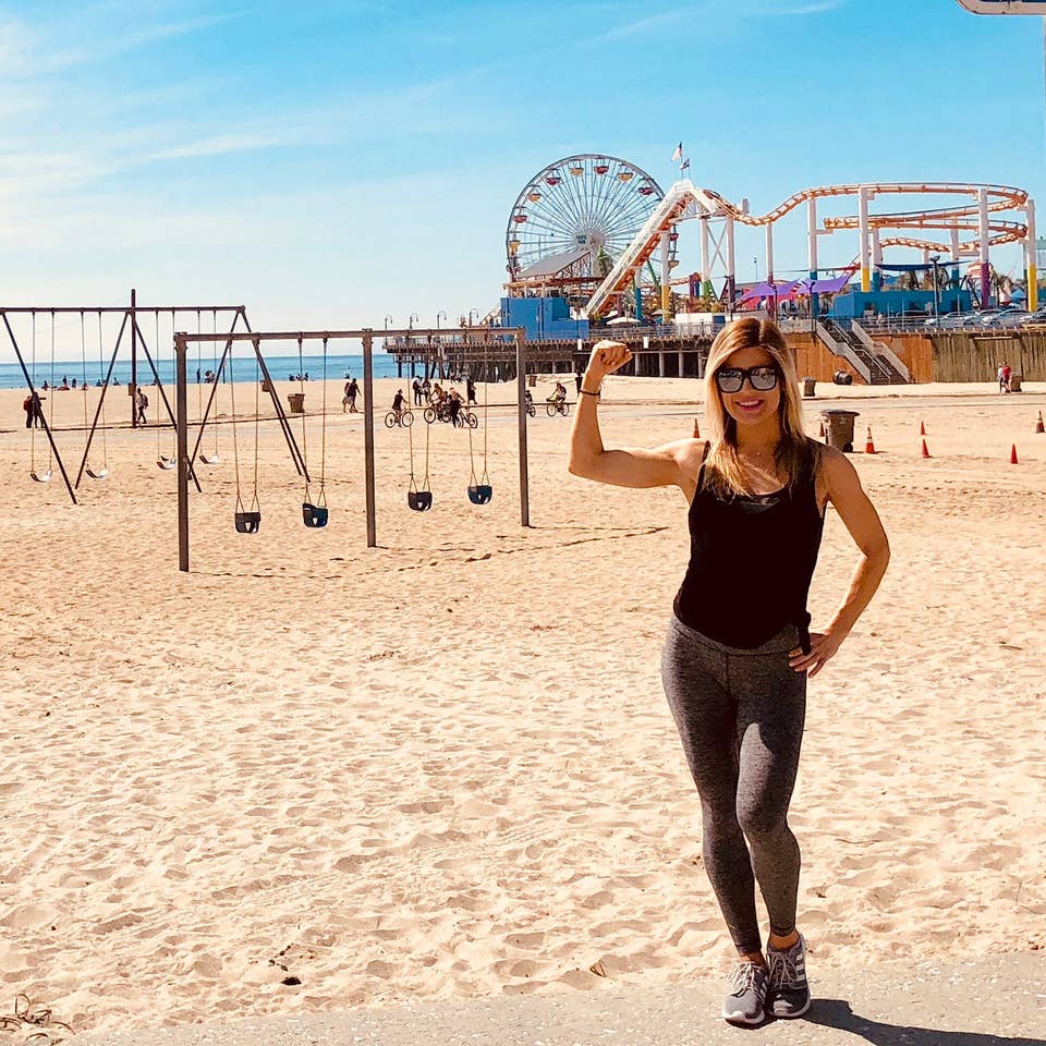 Featured Co-author, Christine, stands on Venice Beach wearing black tanks and leggings in front of the boardwalk and various exercise areas.