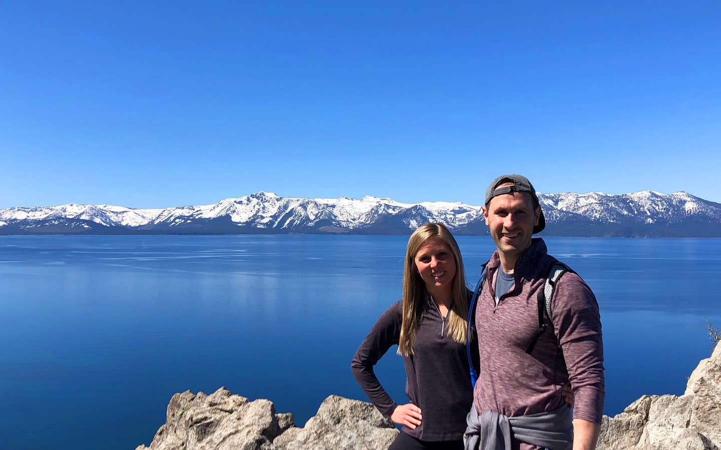 A woman (left) black pullover while hugging a man (right) wearing a red pullover and grey hat in front of a mountain range at Lake Tahoe, Nevada.