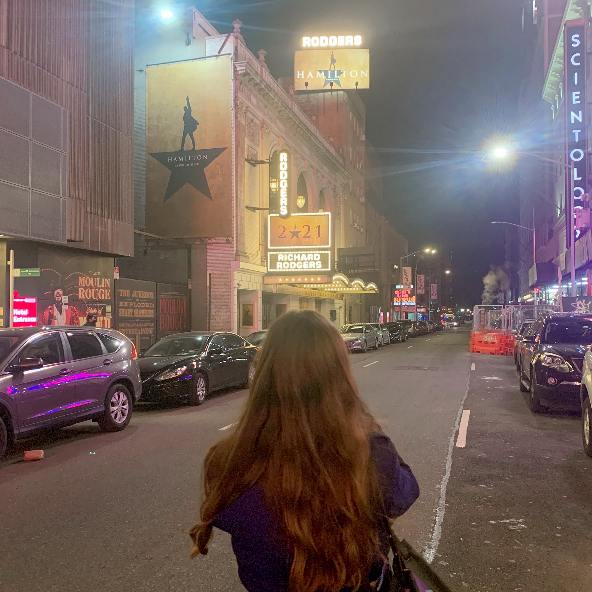 A caucasian woman with long ash-blonde hair wears a jacket under a marquee for the production of 'Hamilton' that reads, '2021' in New York City.