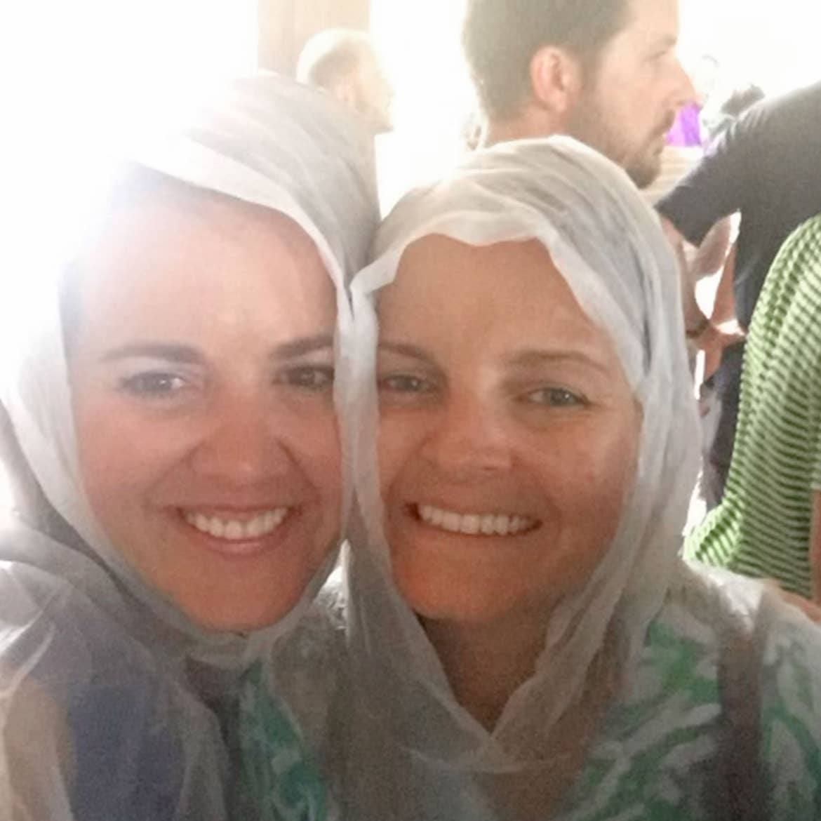 Editor-in-chief, Jenn C. Harmon (left) stands with a friend wearing white ponchos under a roof as it rains in the background.