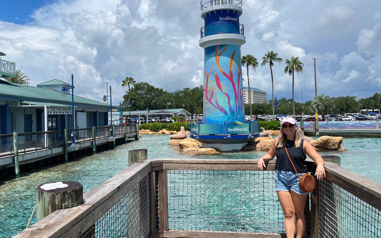 A Caucasian female wearing a pink baseball cap and black tank top stands near the lighthouse in SeaWorld Orlando.