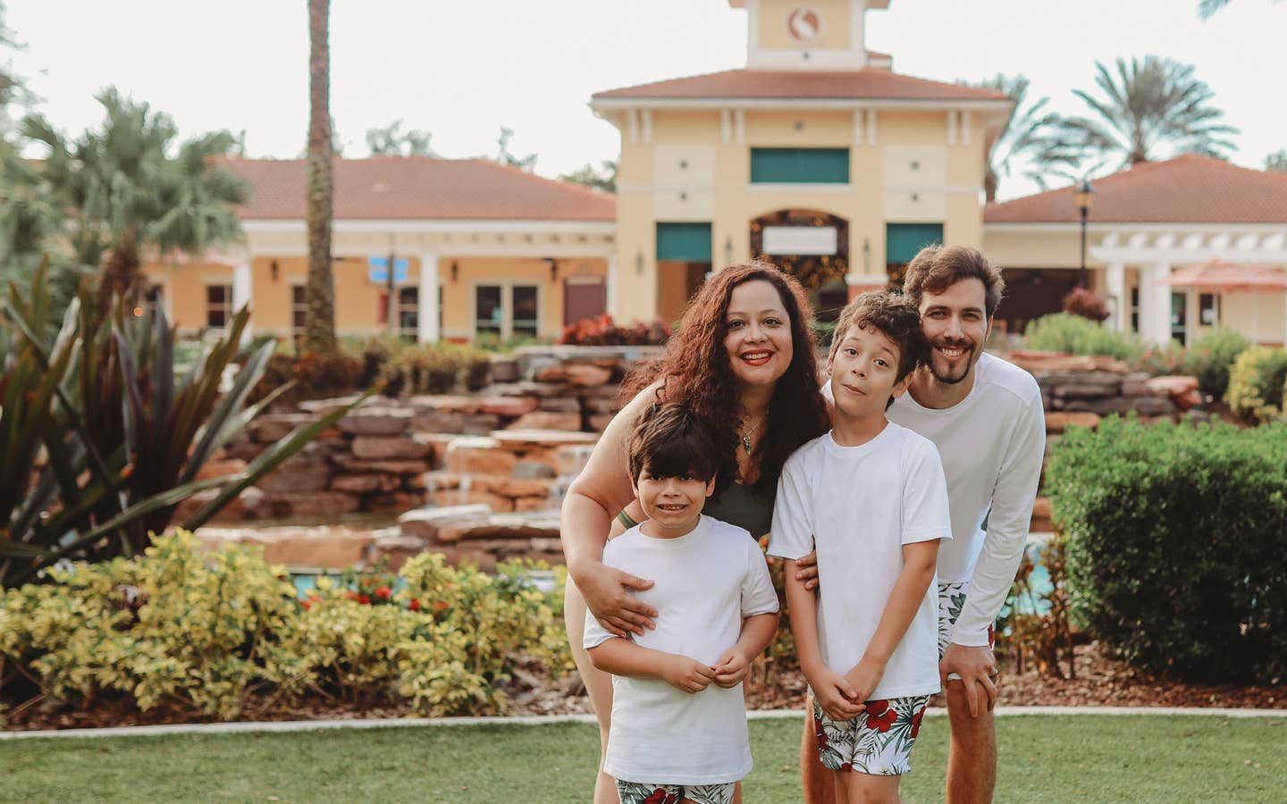 A woman, man and two young boys wear swimwear while standing near a building and lazy river.