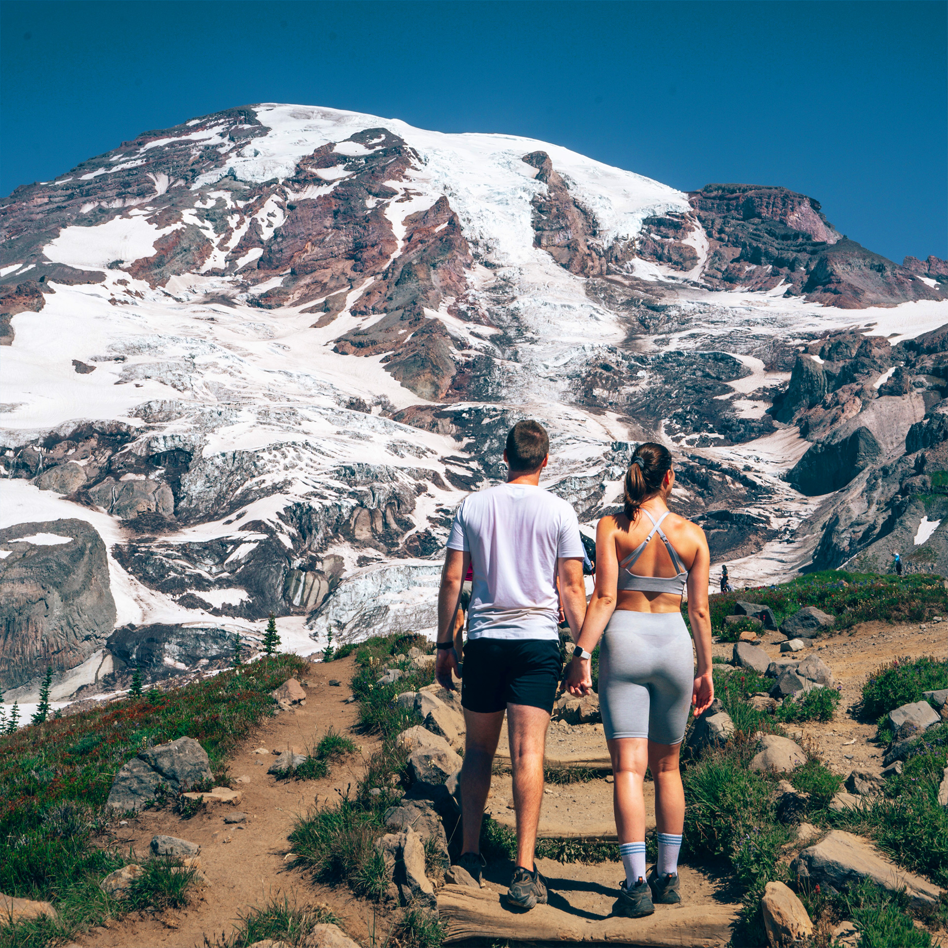 A man in a t-shirt and shorts stands next to a woman in biker shorts and a tank top facing a mountain.