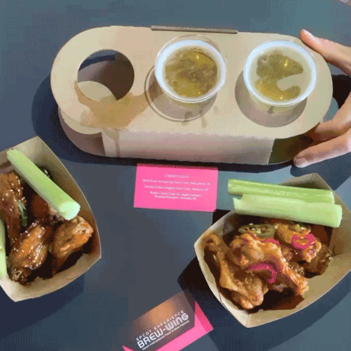 A hand reaches for a small, plastic cup of beer from a cardboard flight container surrounded by hot wings.