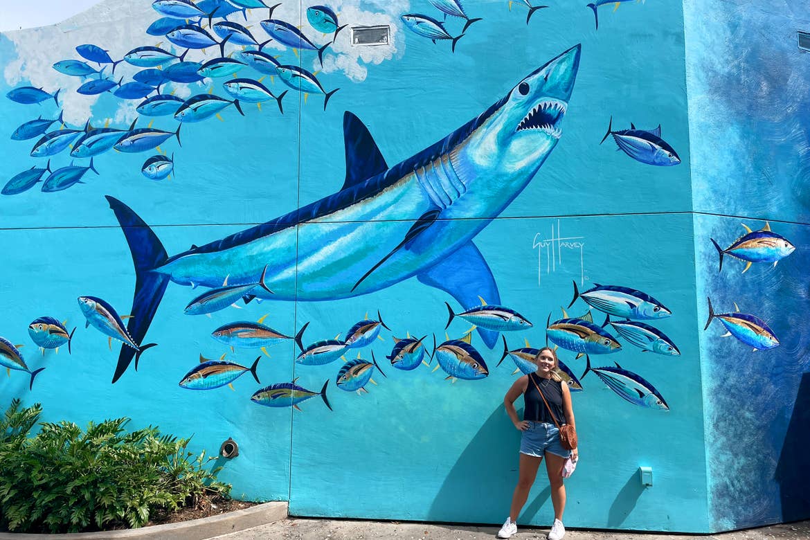 A Caucasian woman with a black tank, denim shorts and white shoes stands next to a mural of a shark surrounded by fish in SeaWorld Orlando.