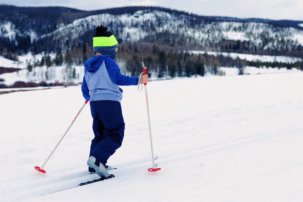 A boy wears cross country ski gear and a multicolor knit hat and blue-grey hoodie in white snow.