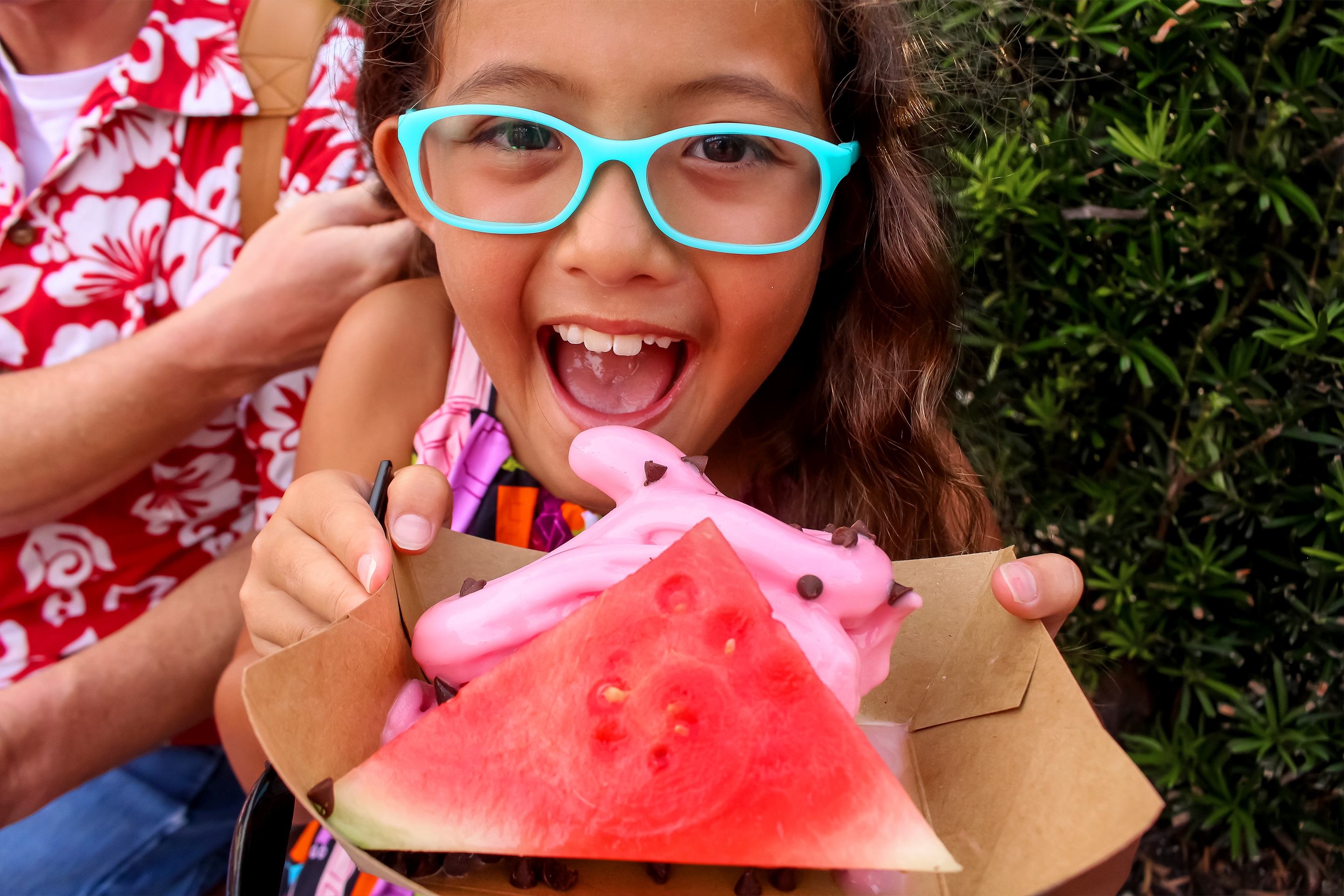 An Asian girl wearing a multi-colored blouse and blue glasses holds a paper container with a slice of watermelon and pink, sot-serve ice cream with mini chocolate chips outdoors.