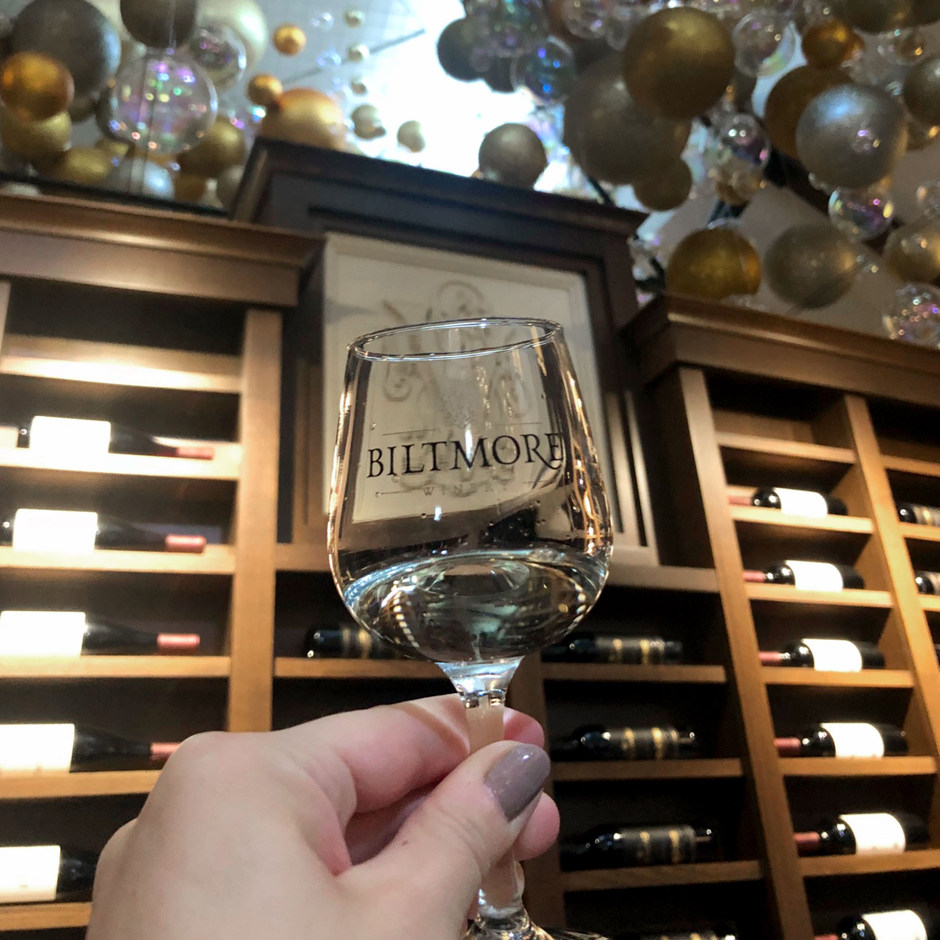 Author, Jennifer C. Harmon, holds a wineglass that reads, 'BILTMORE' in front of a wine shelf, with the ceiling decorated with festive silver, gold and white ornaments.