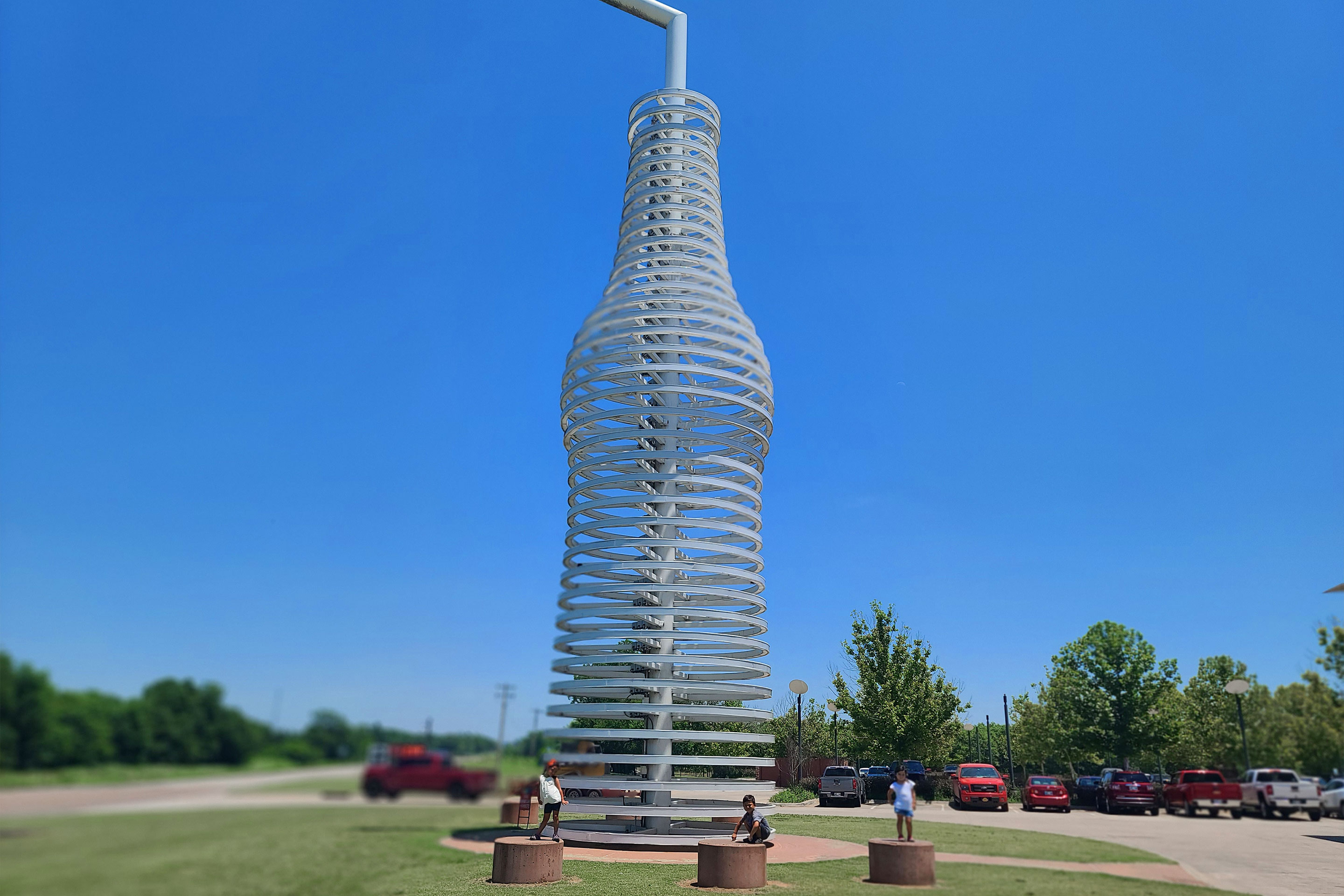 Three toddlers pose on brown, concrete seats under a scaled LED glass soda bottle outdoors.