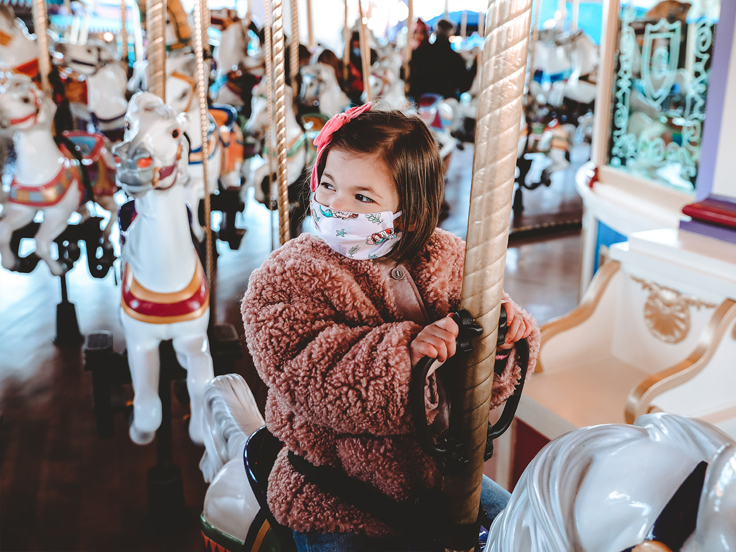 Roux rides a white horse on Prince Charming Regal Carrousel at Magic Kingdom Park at Walt Disney World® Resort wearing a pink fuzzy jacket and a 'Little Mermaid' face mask.