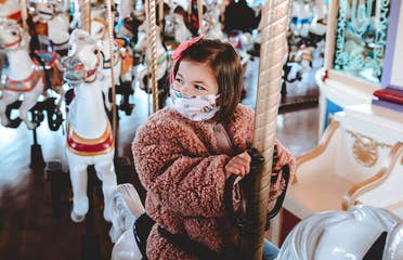Roux rides a white horse on Prince Charming Regal Carrousel at Magic Kingdom Park at Walt Disney World® Resort wearing a pink fuzzy jacket and a 'Little Mermaid' face mask.