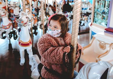 Roux rides a white horse on Prince Charming Regal Carrousel at Magic Kingdom Park at Walt Disney World® Resort wearing a pink fuzzy jacket and a 'Little Mermaid' face mask.
