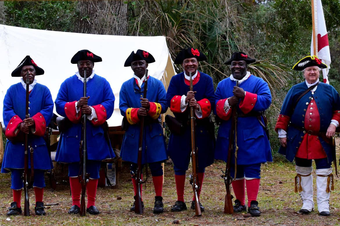 Several African American men and one white man stand in blue, historical war apparel outside near a white tent while holding rifles.