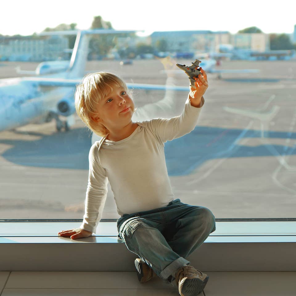 A young boy wearing a white long sleeve shirt plays with a toy airplane in front of an airport window near the runway with a white airplane.
