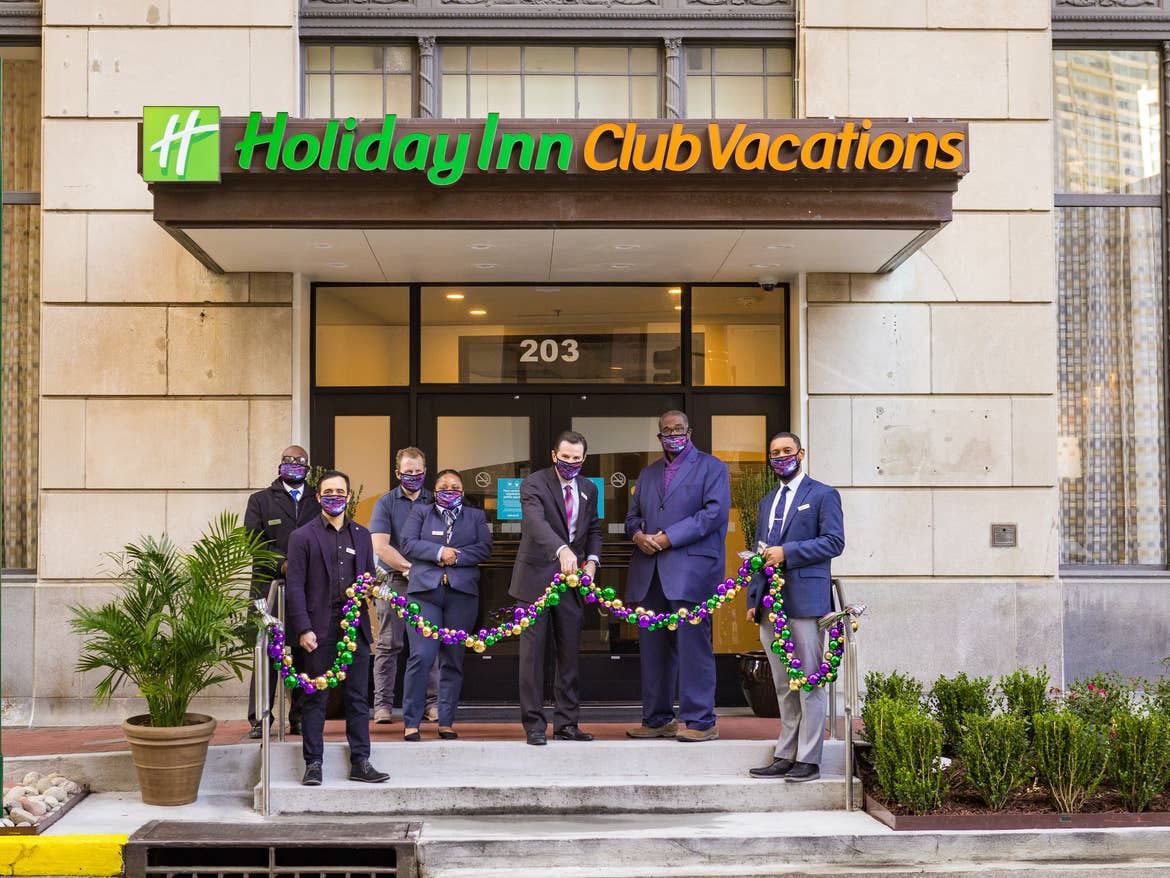 Seven men in suits hold a festive ribbon wreath in front of a building exterior holding ceremonial scissors.