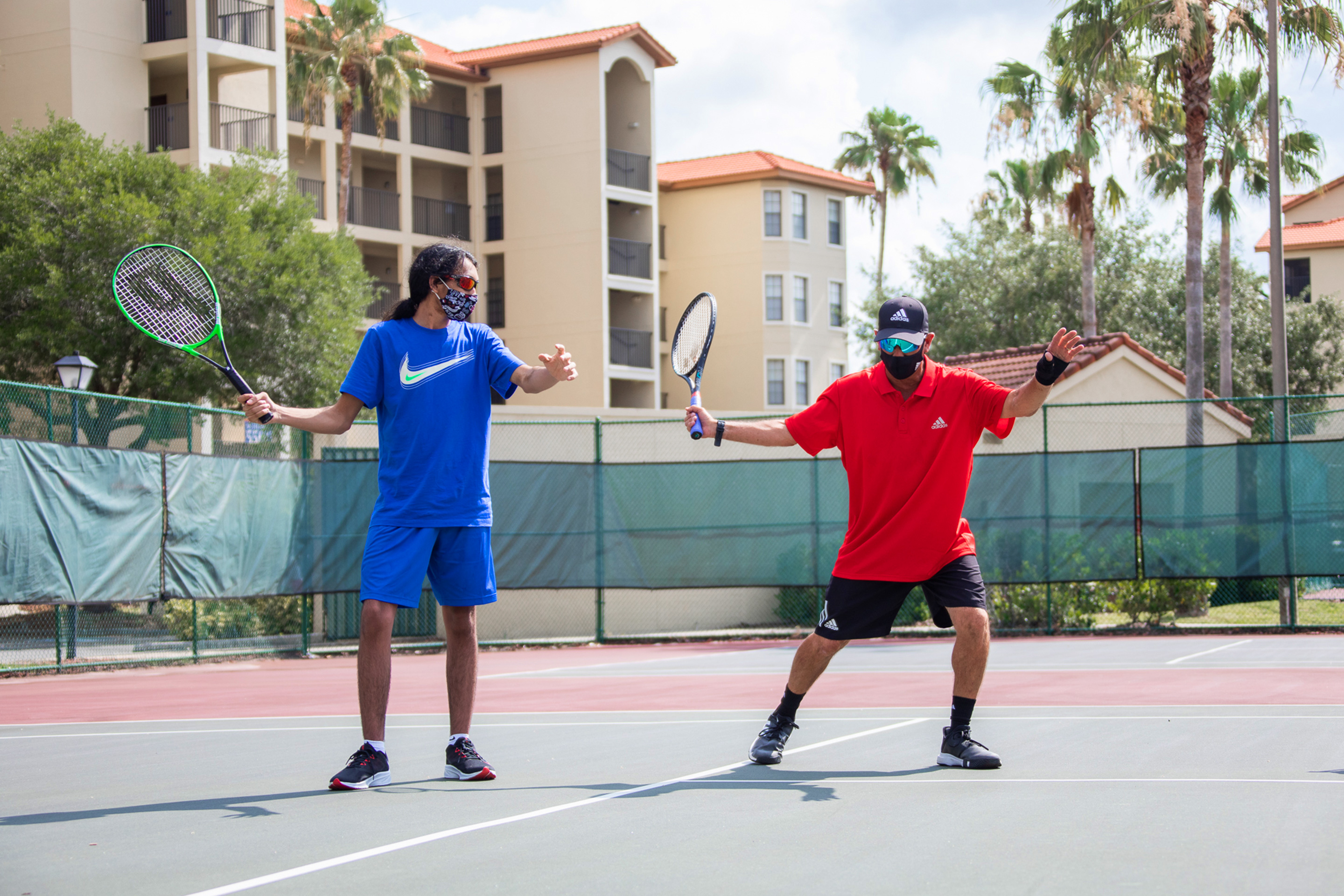 Special Olympic Athlete, Roan Luallen, plays tennis wearing a blue t-shirt and shorts with a safety mask and sunglasses with our instructor, Ernie, on the courts of our Orange Lake Resort located in Orlando, Florida.
